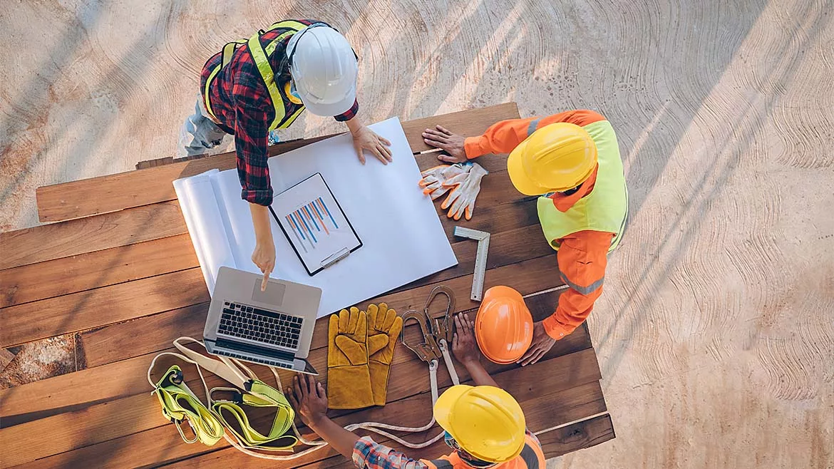 Three construction workers look over a table with blueprints, work equipment, and a laptop