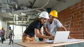 2 people wearing hard hats looking over blueprints and a laptop
