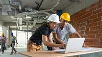 2 people wearing hard hats looking over blueprints and a laptop